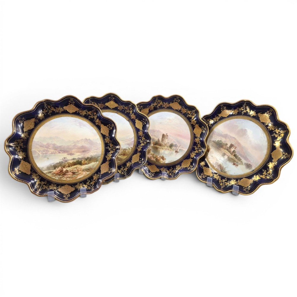 A set of four late nineteenth-century Aynsley porcelain cabinet plates featuring hand-painted landscape scenes surrounded by cobalt blue rims with heavy gold foliage gilding.