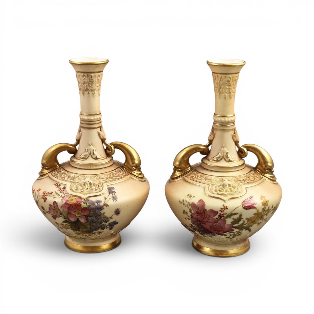 A symmetrical pair of Edwardian Royal Worcester blush ivory porcelain vases featuring gilded dolphin handles, relief-moulded necks, and hand-painted floral decoration.