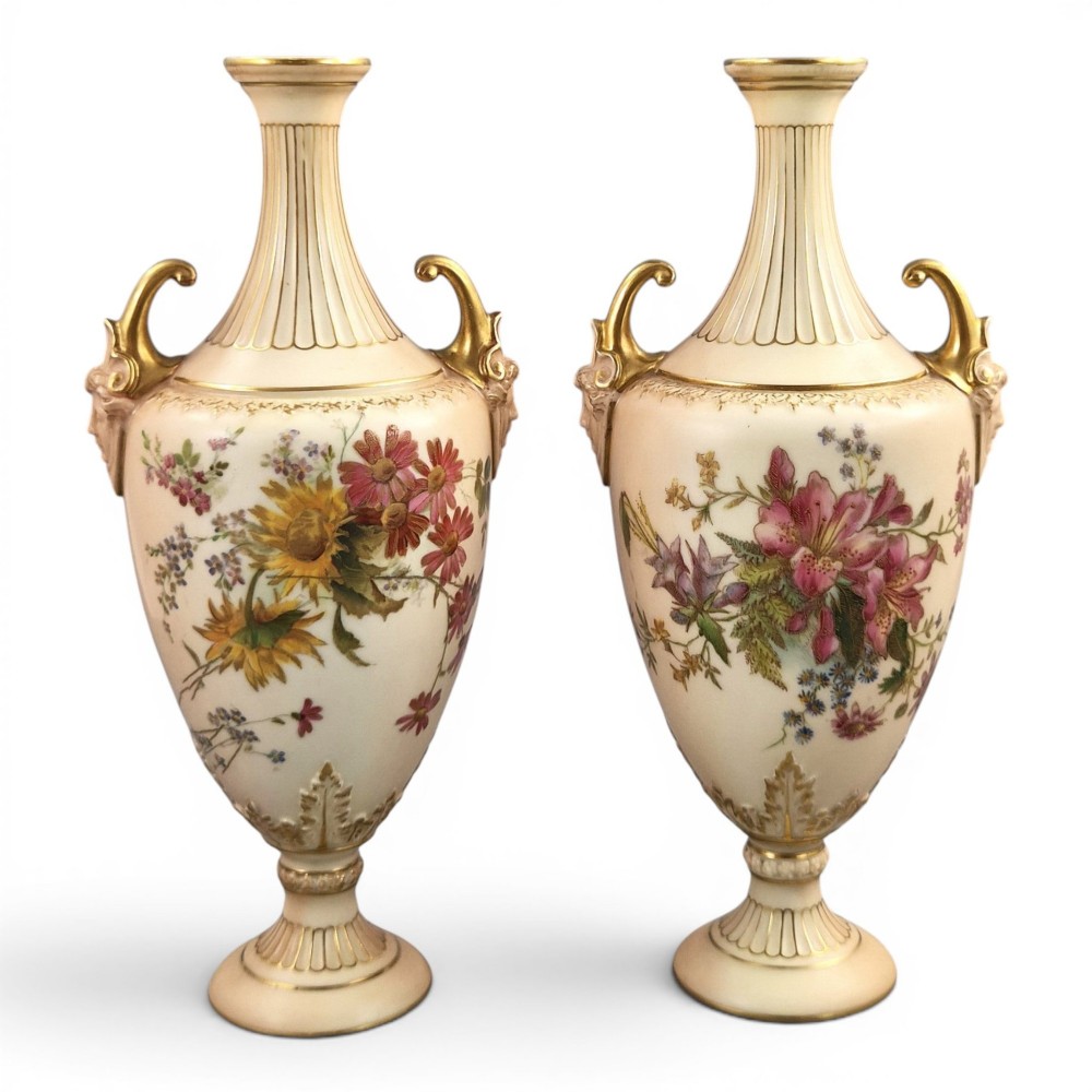 A pair of Royal Worcester blush ivory porcelain urn-shaped vases featuring hand-painted floral motifs, gold-gilded handles, fluted necks, and circular pedestal bases.