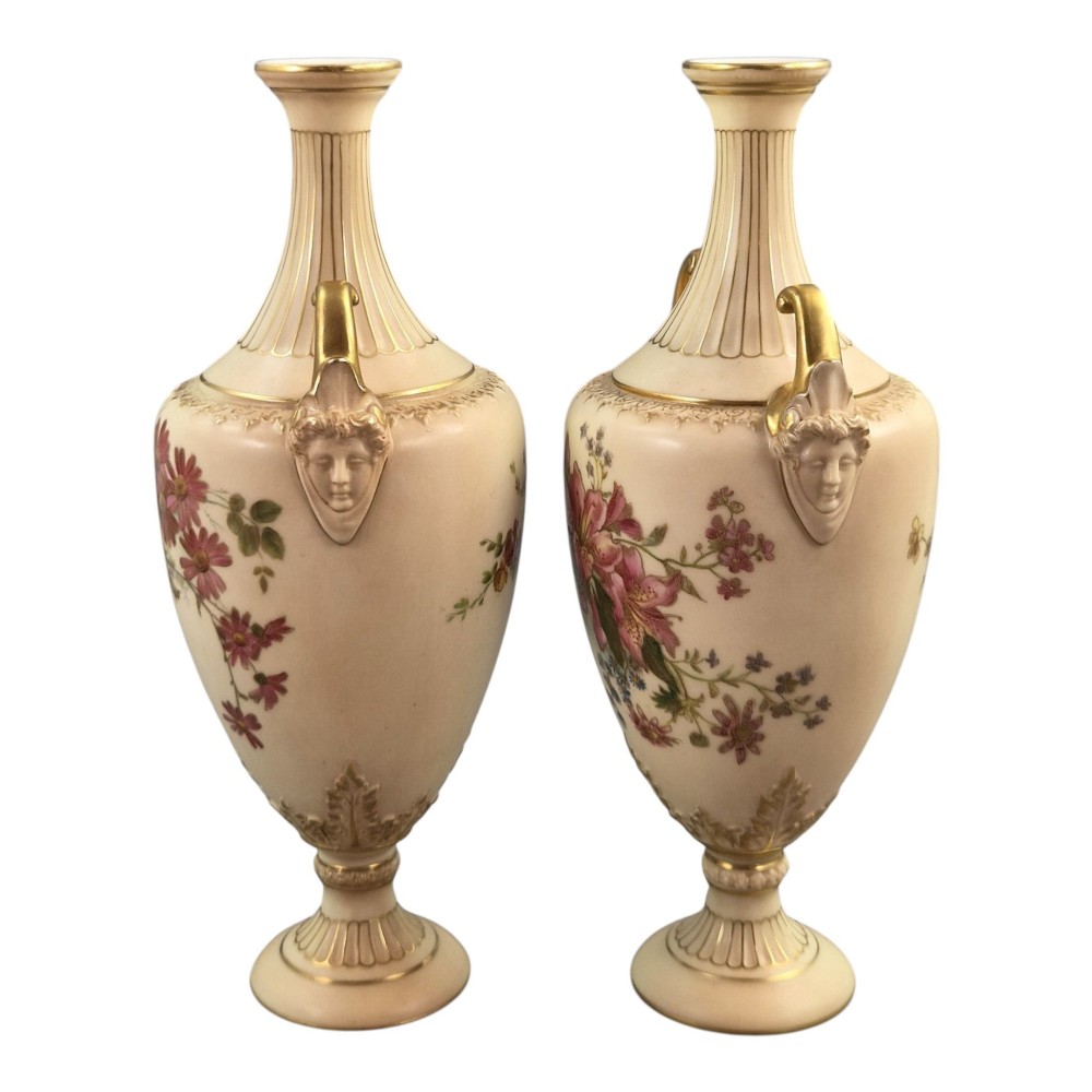 A pair of Royal Worcester blush ivory porcelain urn-shaped vases featuring hand-painted floral motifs, gold-gilded handles, fluted necks, and circular pedestal bases. View II