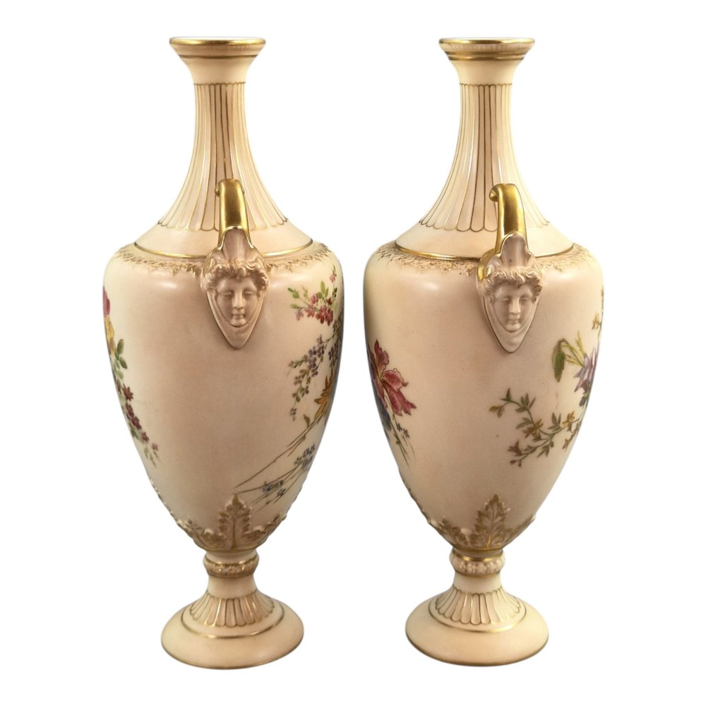 A pair of Royal Worcester blush ivory porcelain urn-shaped vases featuring hand-painted floral motifs, gold-gilded handles, fluted necks, and circular pedestal bases. View IV