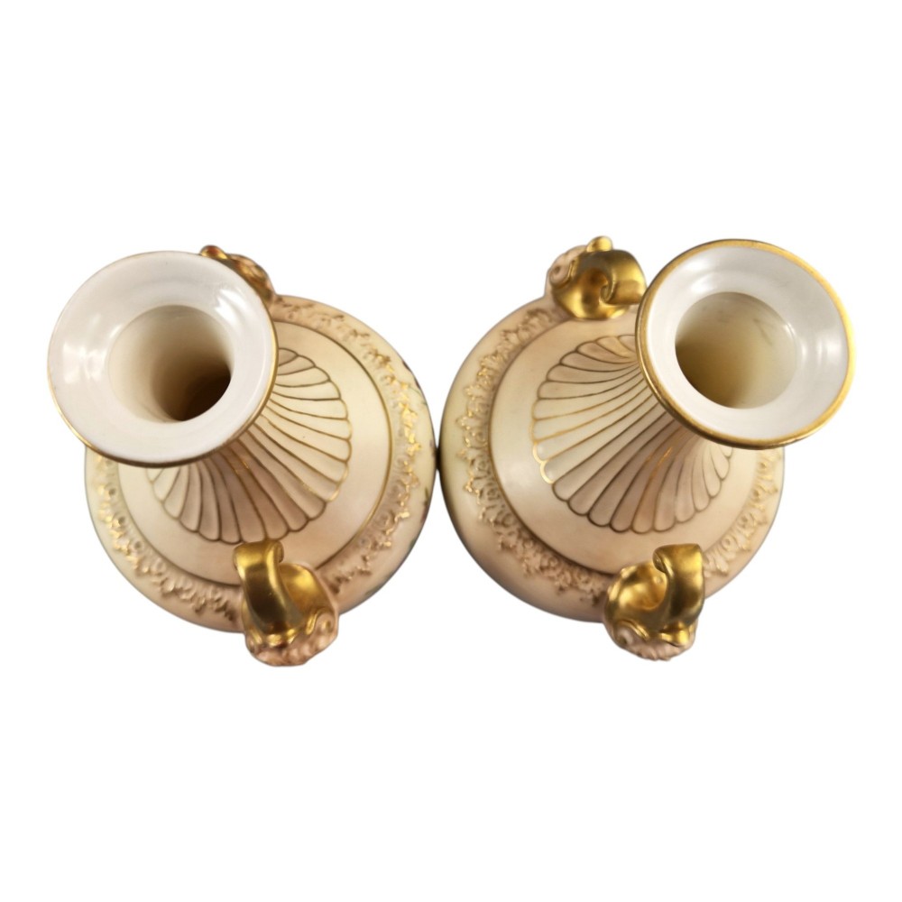 A pair of Royal Worcester blush ivory porcelain urn-shaped vases featuring hand-painted floral motifs, gold-gilded handles, fluted necks, and circular pedestal bases. View V