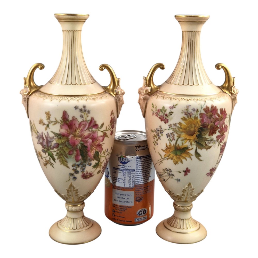 A pair of Royal Worcester blush ivory porcelain urn-shaped vases featuring hand-painted floral motifs, gold-gilded handles, fluted necks, and circular pedestal bases. View XII