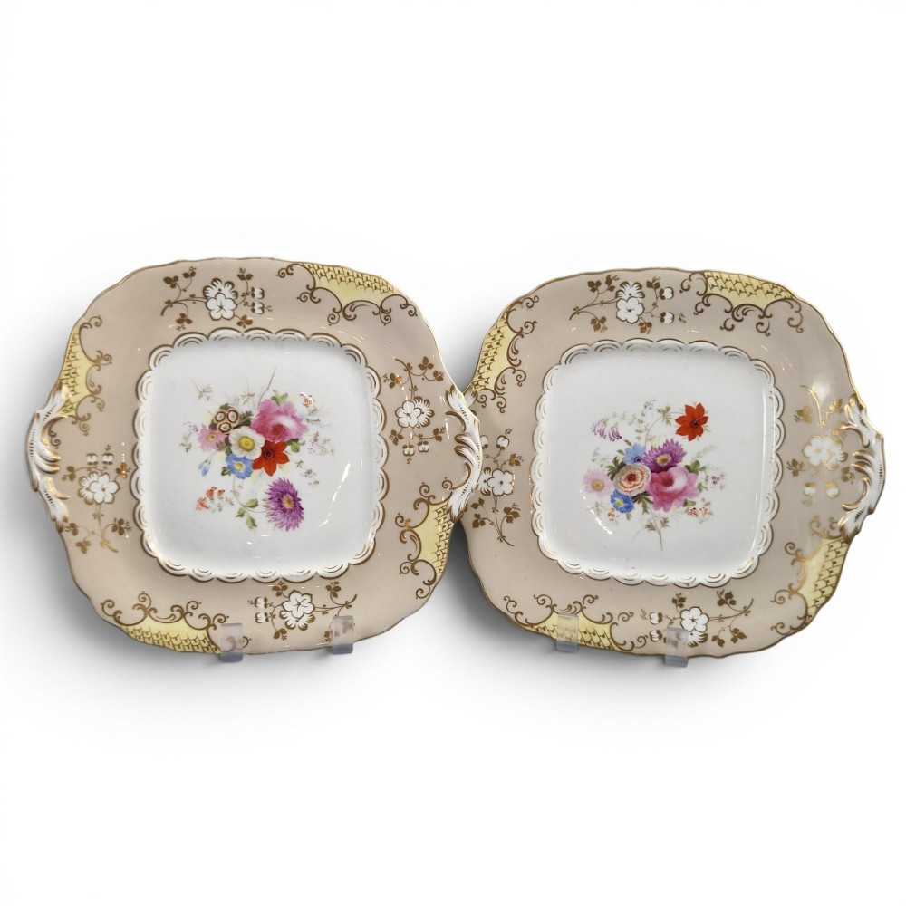 A pair of early Victorian Coalport porcelain square dessert dishes featuring hand-painted floral centres, beige borders with gold scrollwork, and moulded twin handles.