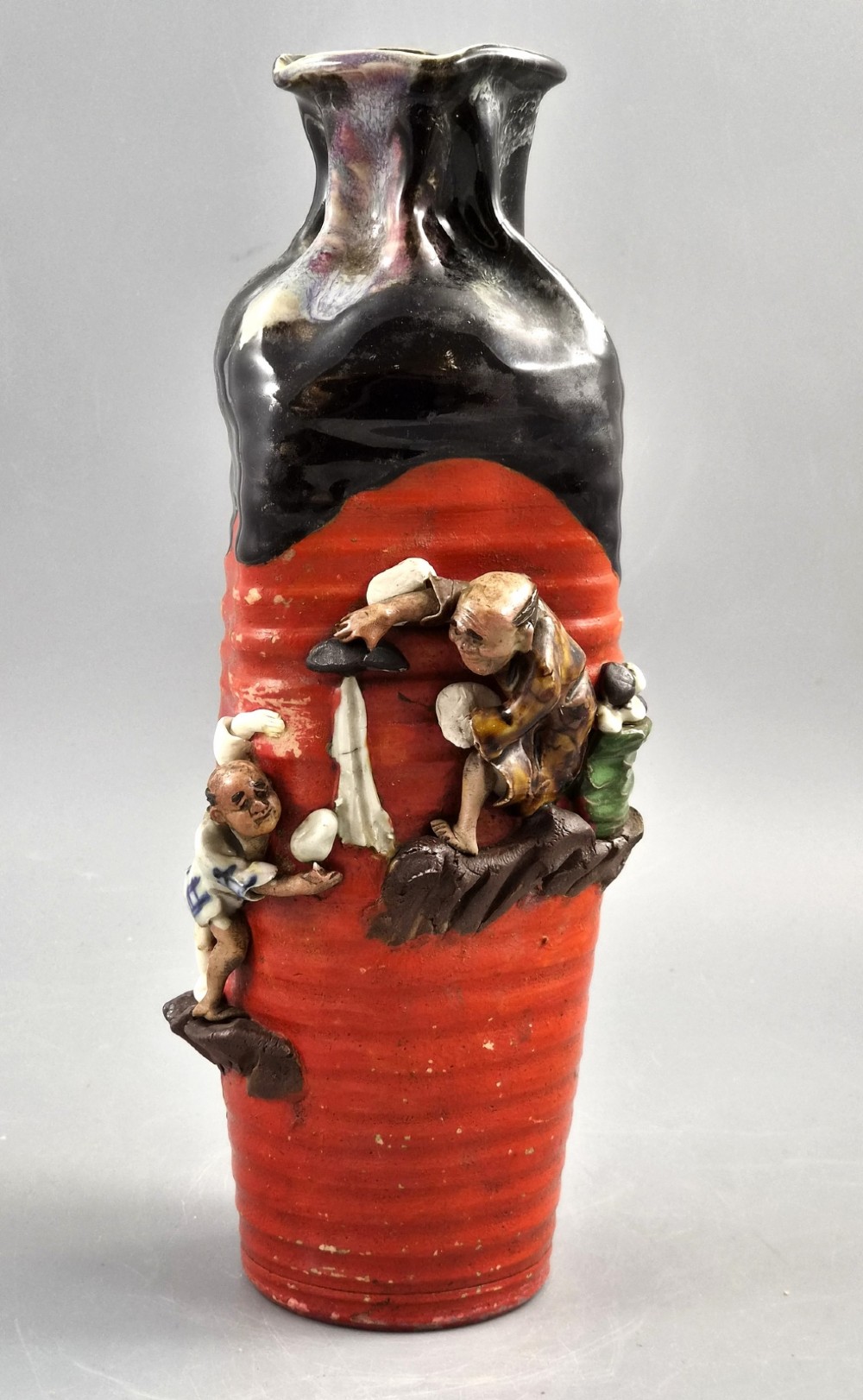 A tall Japanese Sumida Gawa ceramic vase featuring a ribbed red base, applied relief figures of sages on a cliff, and a heavy dark drip glaze around the rim.
