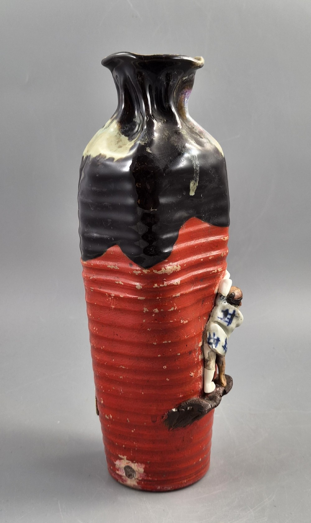 A tall Japanese Sumida Gawa ceramic vase featuring a ribbed red base, applied relief figures of sages on a cliff, and a heavy dark drip glaze around the rim. View V