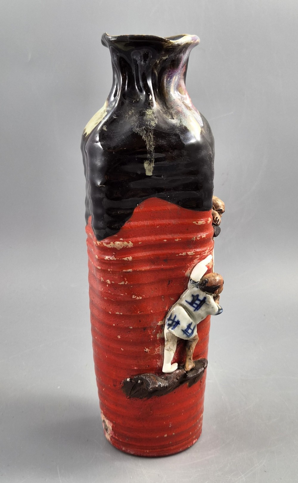 A tall Japanese Sumida Gawa ceramic vase featuring a ribbed red base, applied relief figures of sages on a cliff, and a heavy dark drip glaze around the rim. View VI