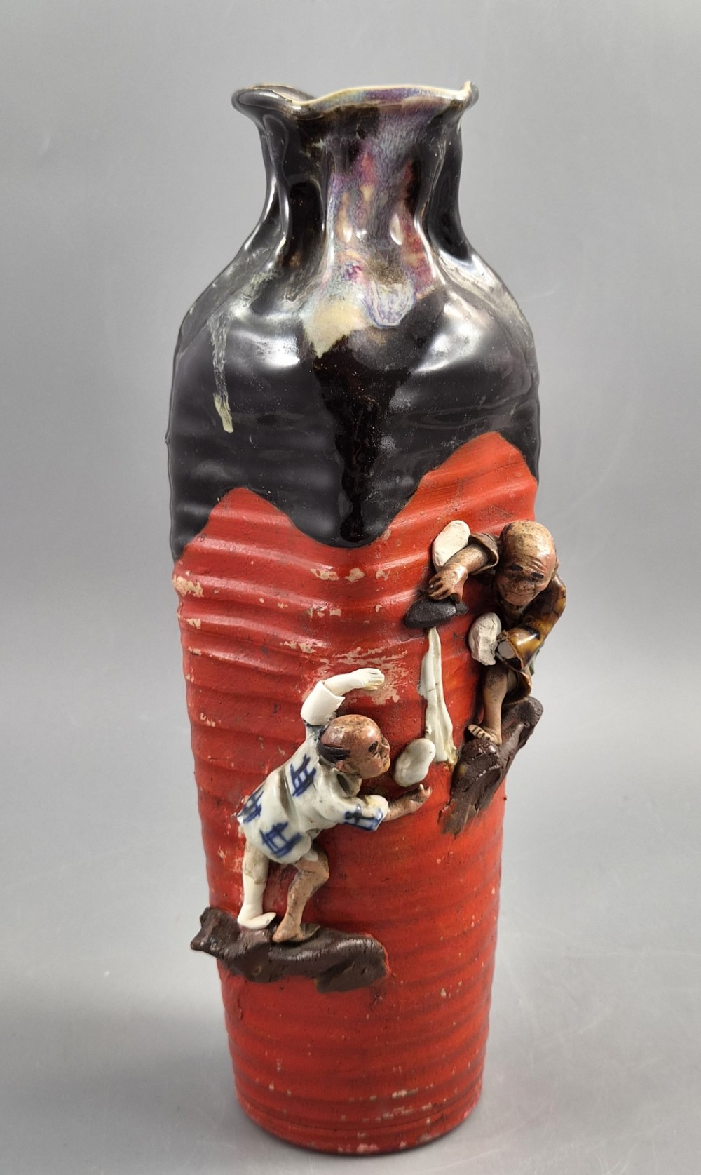 A tall Japanese Sumida Gawa ceramic vase featuring a ribbed red base, applied relief figures of sages on a cliff, and a heavy dark drip glaze around the rim. View VII