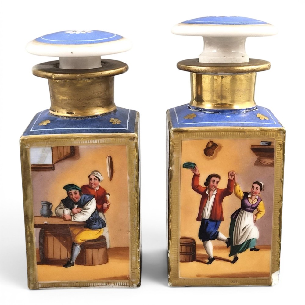 A pair of square 19th-century French porcelain perfume bottles featuring cobalt blue tops, gilded necks, and hand-painted genre scenes of figures in tavern settings.