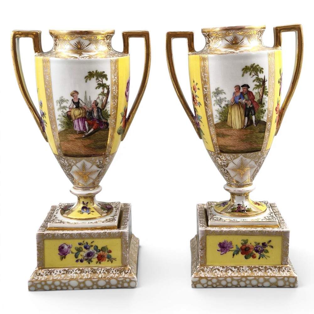 A pair of late nineteenth-century Dresden porcelain urns featuring yellow ground, hand-painted romantic scenes, gilded handles, and square pedestal bases.