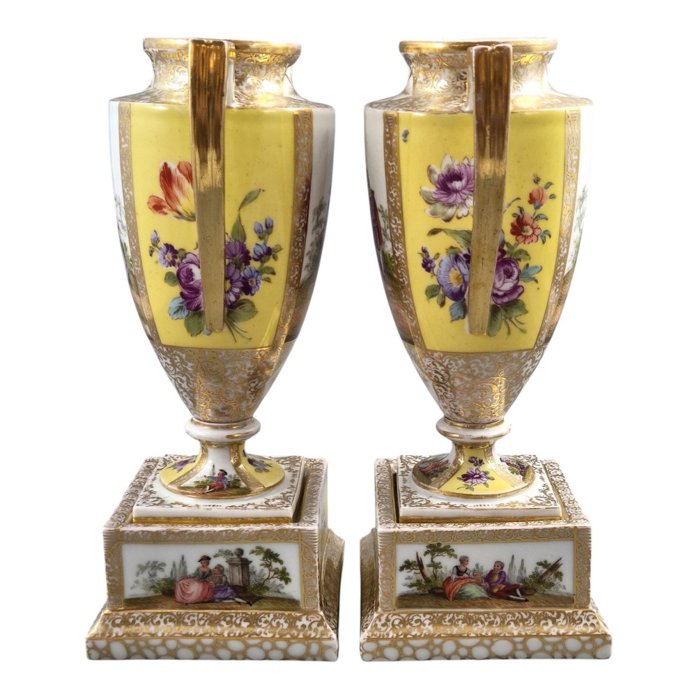 A pair of late nineteenth-century Dresden porcelain urns featuring yellow ground, hand-painted romantic scenes, gilded handles, and square pedestal bases. View IV
