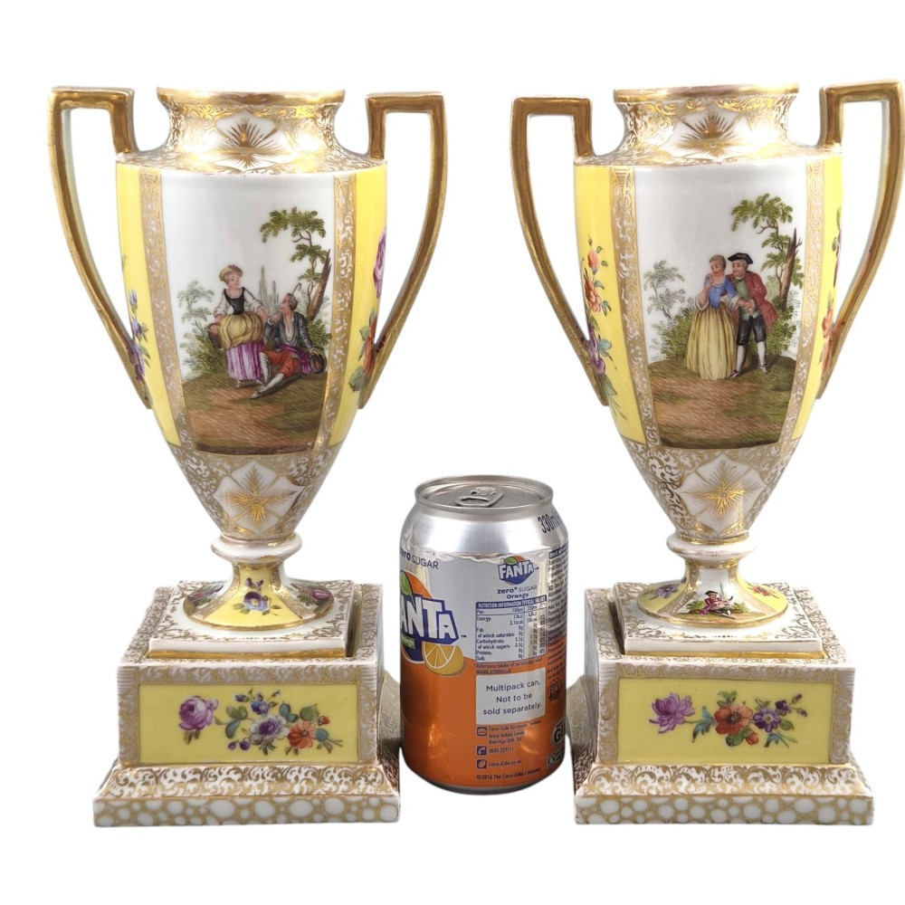 A pair of late nineteenth-century Dresden porcelain urns featuring yellow ground, hand-painted romantic scenes, gilded handles, and square pedestal bases. View V