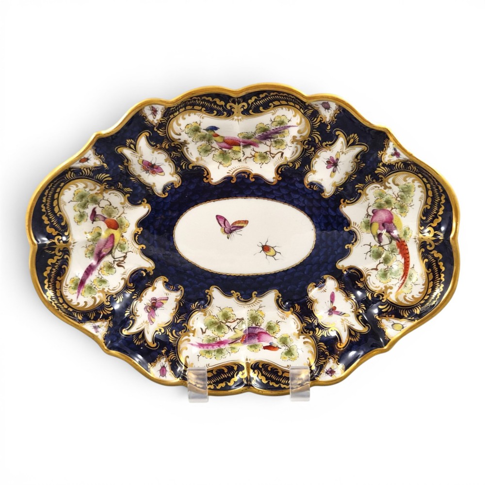 An oval scalloped porcelain dessert dish featuring a deep blue scale ground pattern with gilt-framed reserves containing hand-painted exotic birds and insects.
