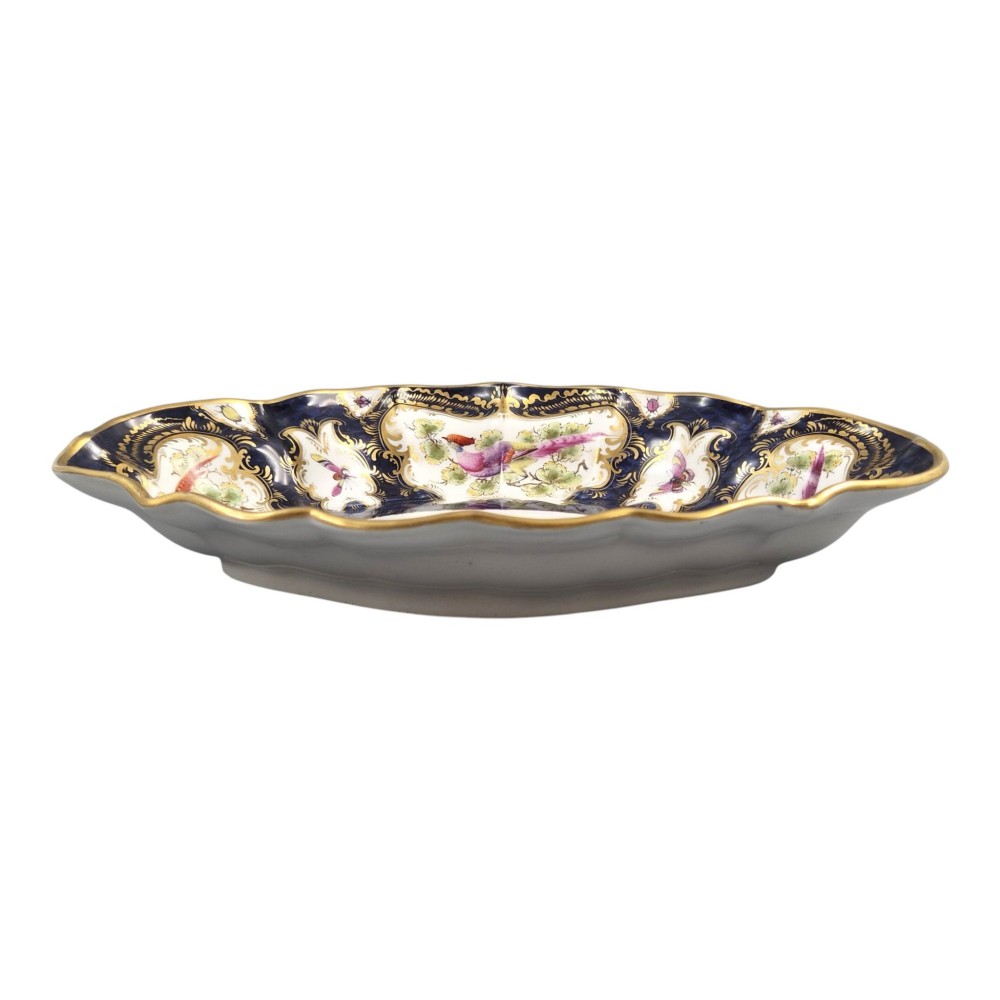 An oval scalloped porcelain dessert dish featuring a deep blue scale ground pattern with gilt-framed reserves containing hand-painted exotic birds and insects. View VI