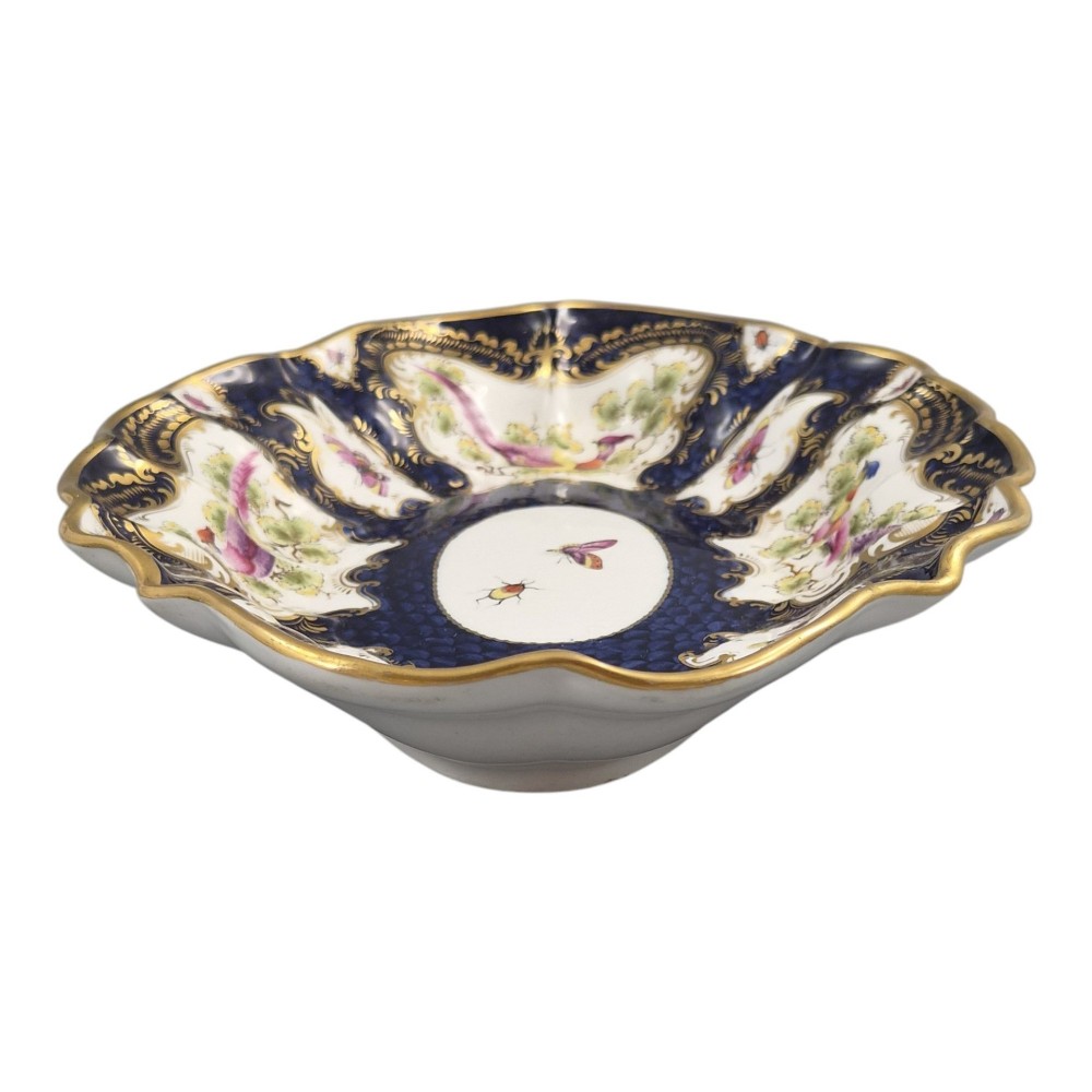 An oval scalloped porcelain dessert dish featuring a deep blue scale ground pattern with gilt-framed reserves containing hand-painted exotic birds and insects. View IX