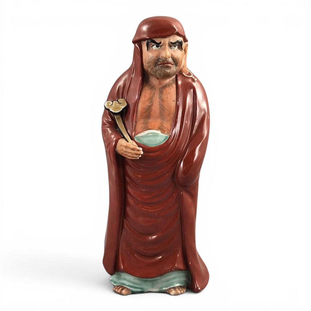 A standing Japanese Kutani porcelain okimono figure depicting Bodhidharma wearing draped terracotta red robes and holding a sceptre.
