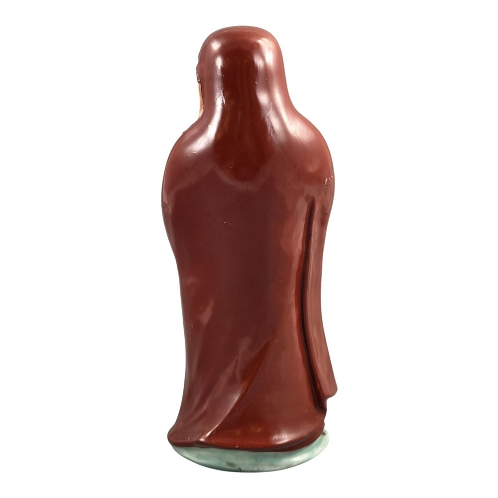 A standing Japanese Kutani porcelain okimono figure depicting Bodhidharma wearing draped terracotta red robes and holding a sceptre. View IV