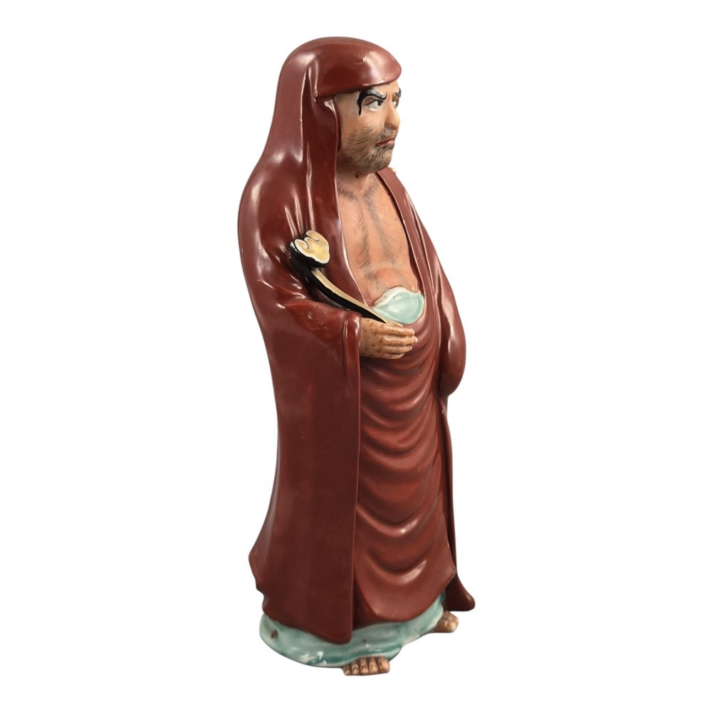 A standing Japanese Kutani porcelain okimono figure depicting Bodhidharma wearing draped terracotta red robes and holding a sceptre. View VI