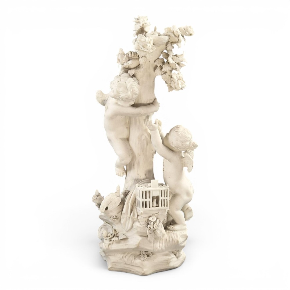 An unglazed off-white biscuit porcelain figure depicting two winged cherubs beside a tree trunk with a birdcage near the base.