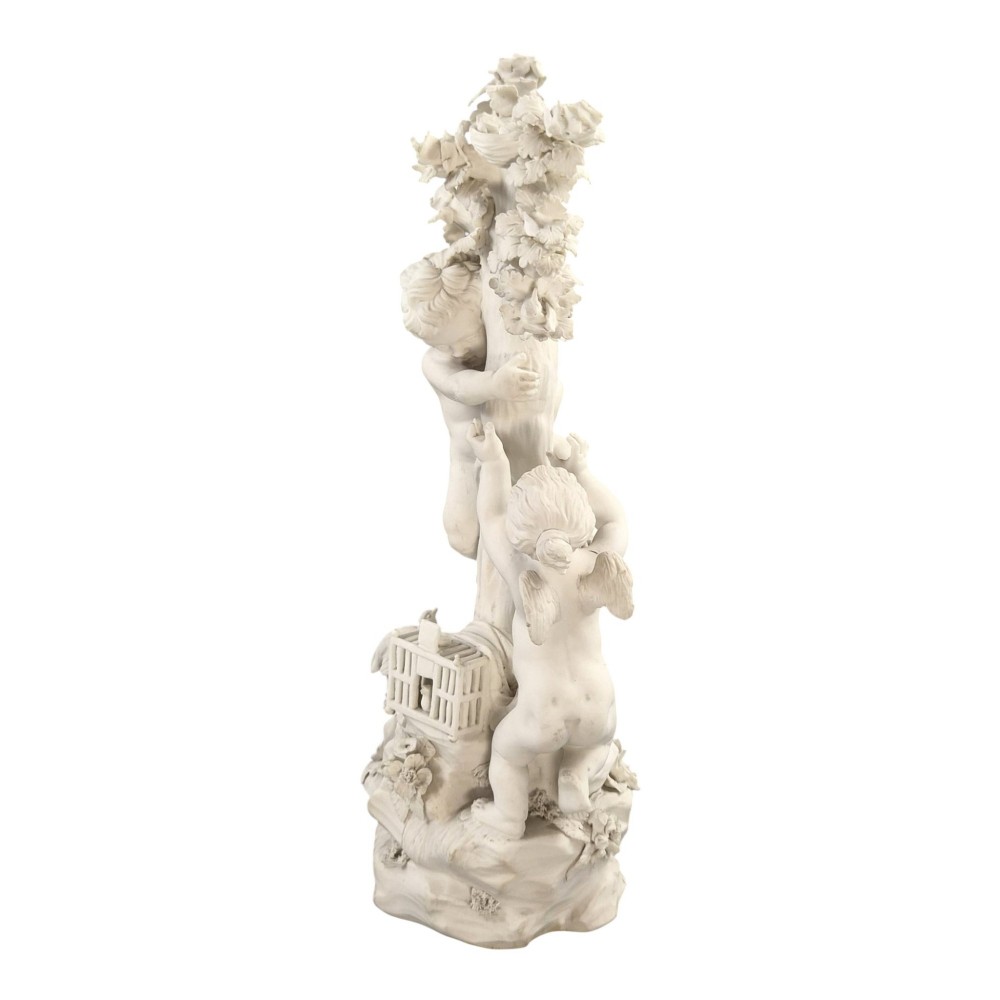 An unglazed off-white biscuit porcelain figure depicting two winged cherubs beside a tree trunk with a birdcage near the base. View II
