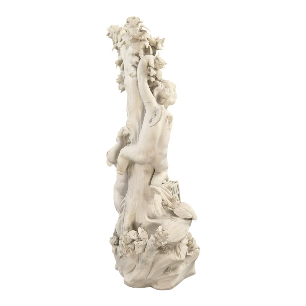 An unglazed off-white biscuit porcelain figure depicting two winged cherubs beside a tree trunk with a birdcage near the base. View V