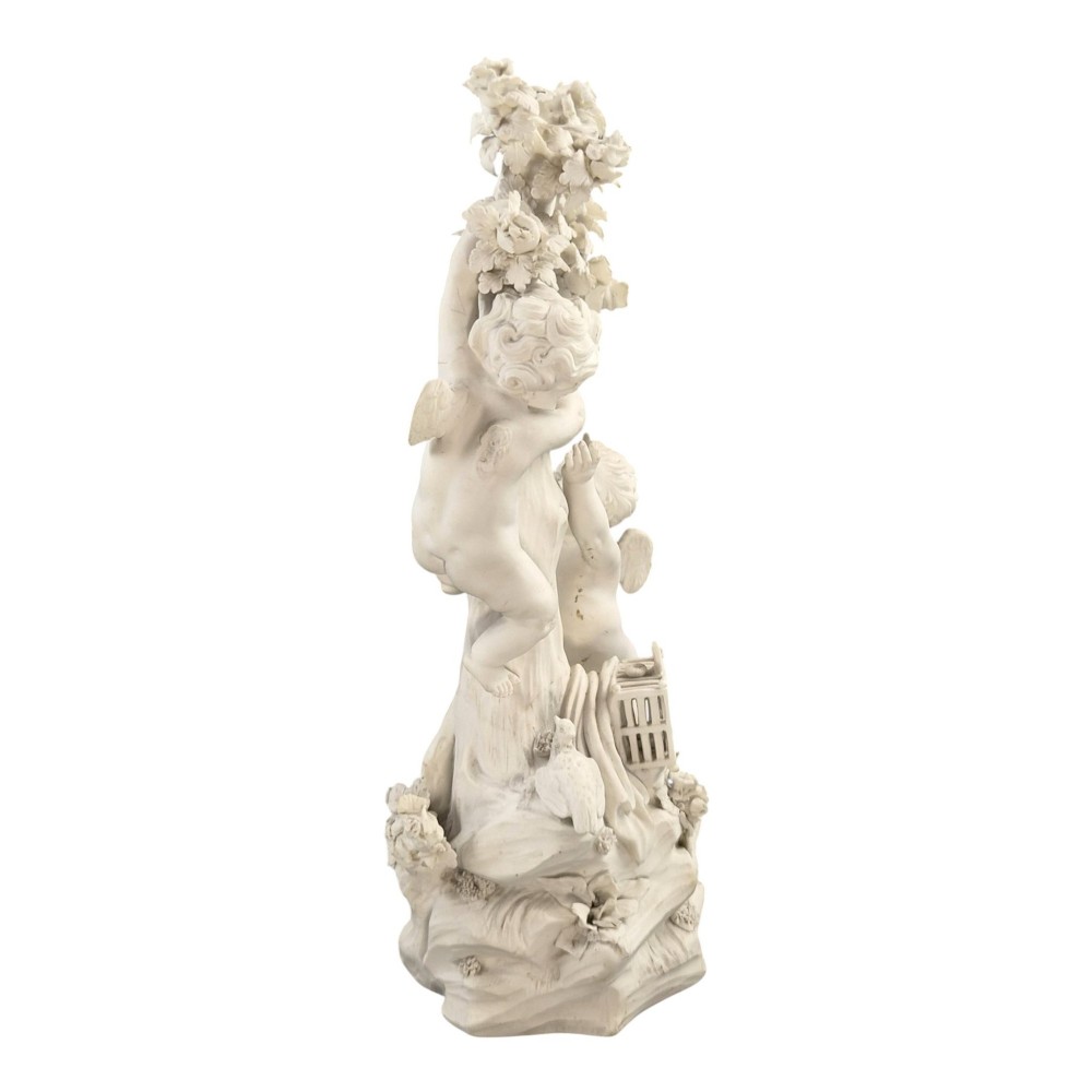 An unglazed off-white biscuit porcelain figure depicting two winged cherubs beside a tree trunk with a birdcage near the base. View VI