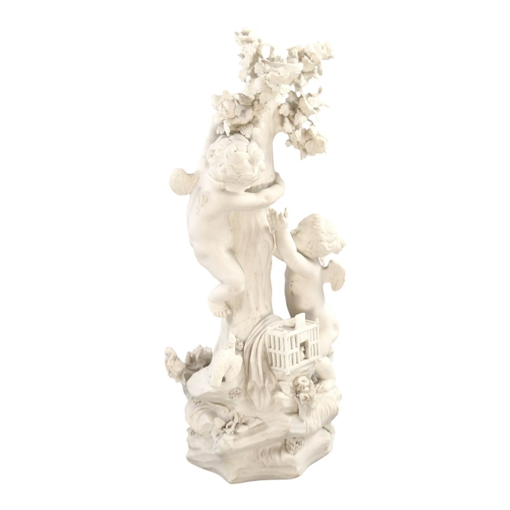 An unglazed off-white biscuit porcelain figure depicting two winged cherubs beside a tree trunk with a birdcage near the base. View VII