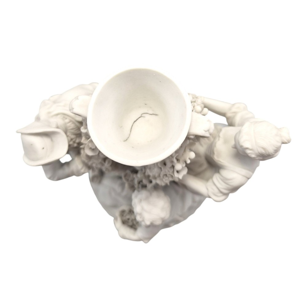 A white matte biscuit porcelain sculptural group depicting three figures and a central urn adorned with delicate floral garlands on a decorative circular base. View III