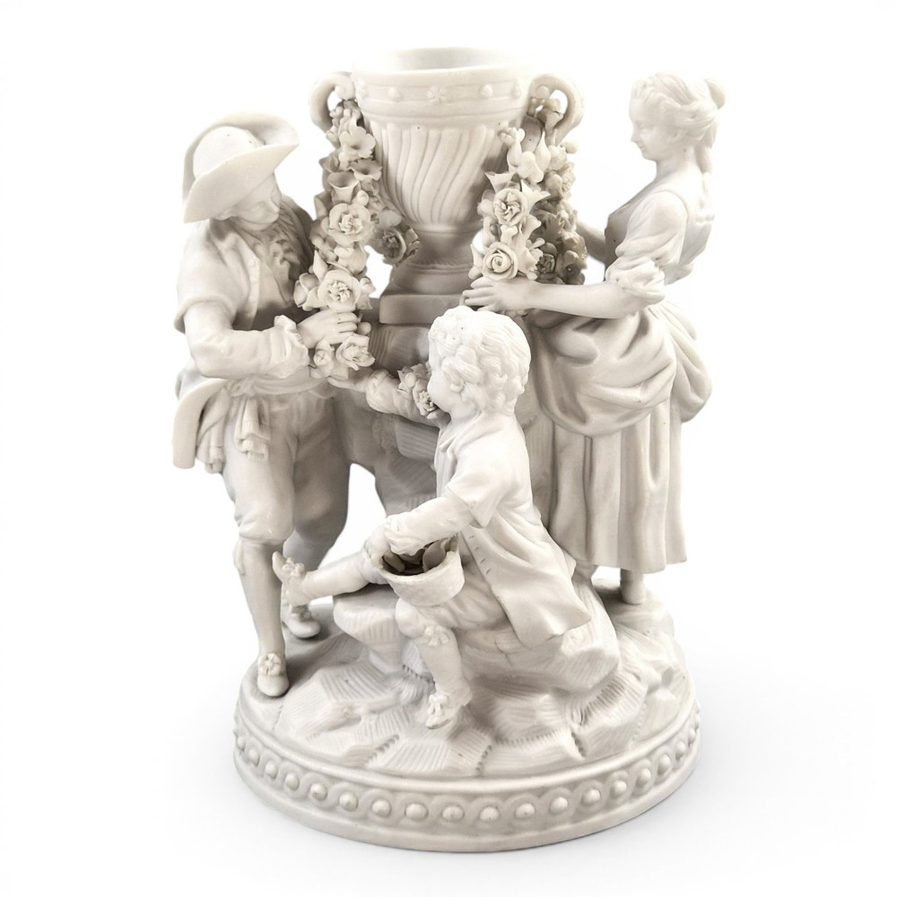 A white matte biscuit porcelain sculptural group depicting three figures and a central urn adorned with delicate floral garlands on a decorative circular base.