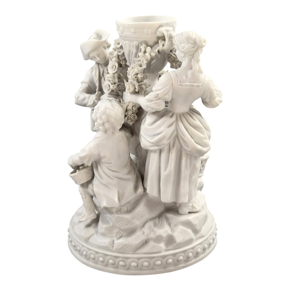 A white matte biscuit porcelain sculptural group depicting three figures and a central urn adorned with delicate floral garlands on a decorative circular base. View IV