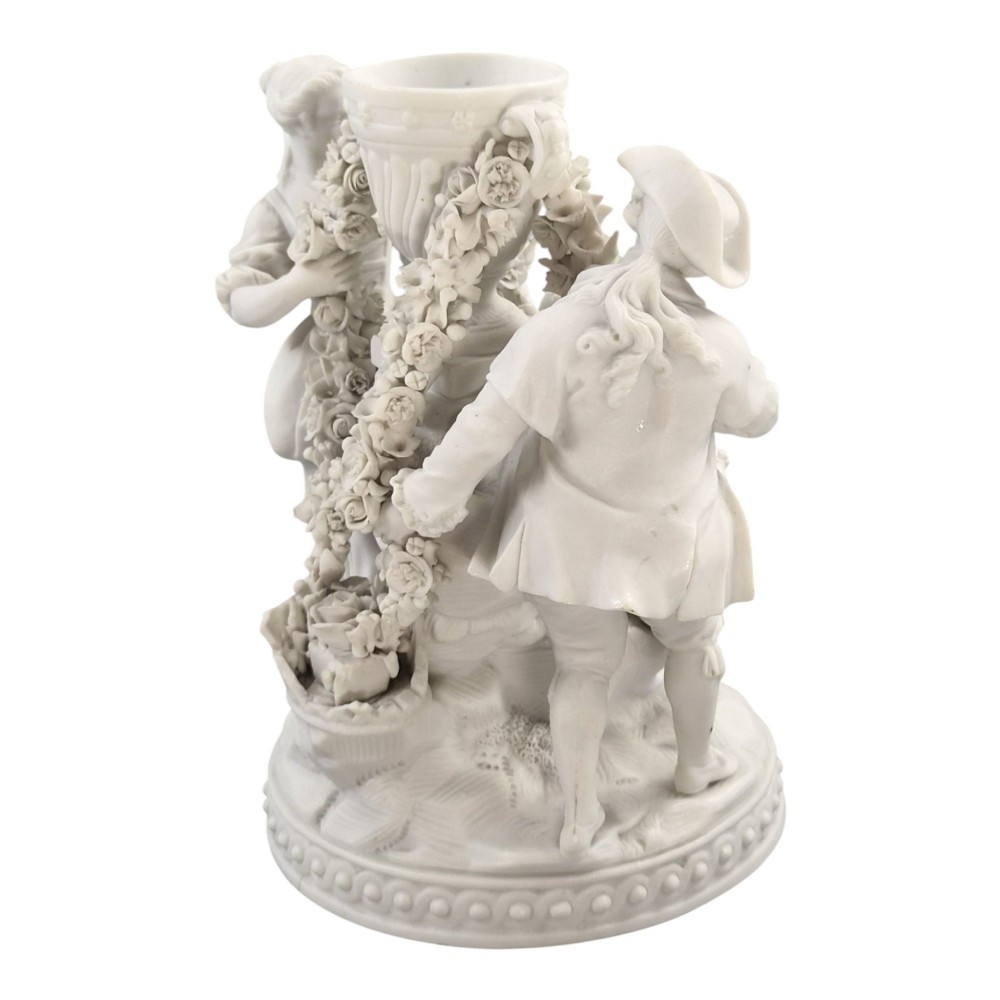 A white matte biscuit porcelain sculptural group depicting three figures and a central urn adorned with delicate floral garlands on a decorative circular base. View VII