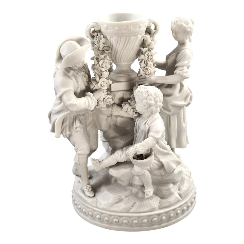 A white matte biscuit porcelain sculptural group depicting three figures and a central urn adorned with delicate floral garlands on a decorative circular base. View X