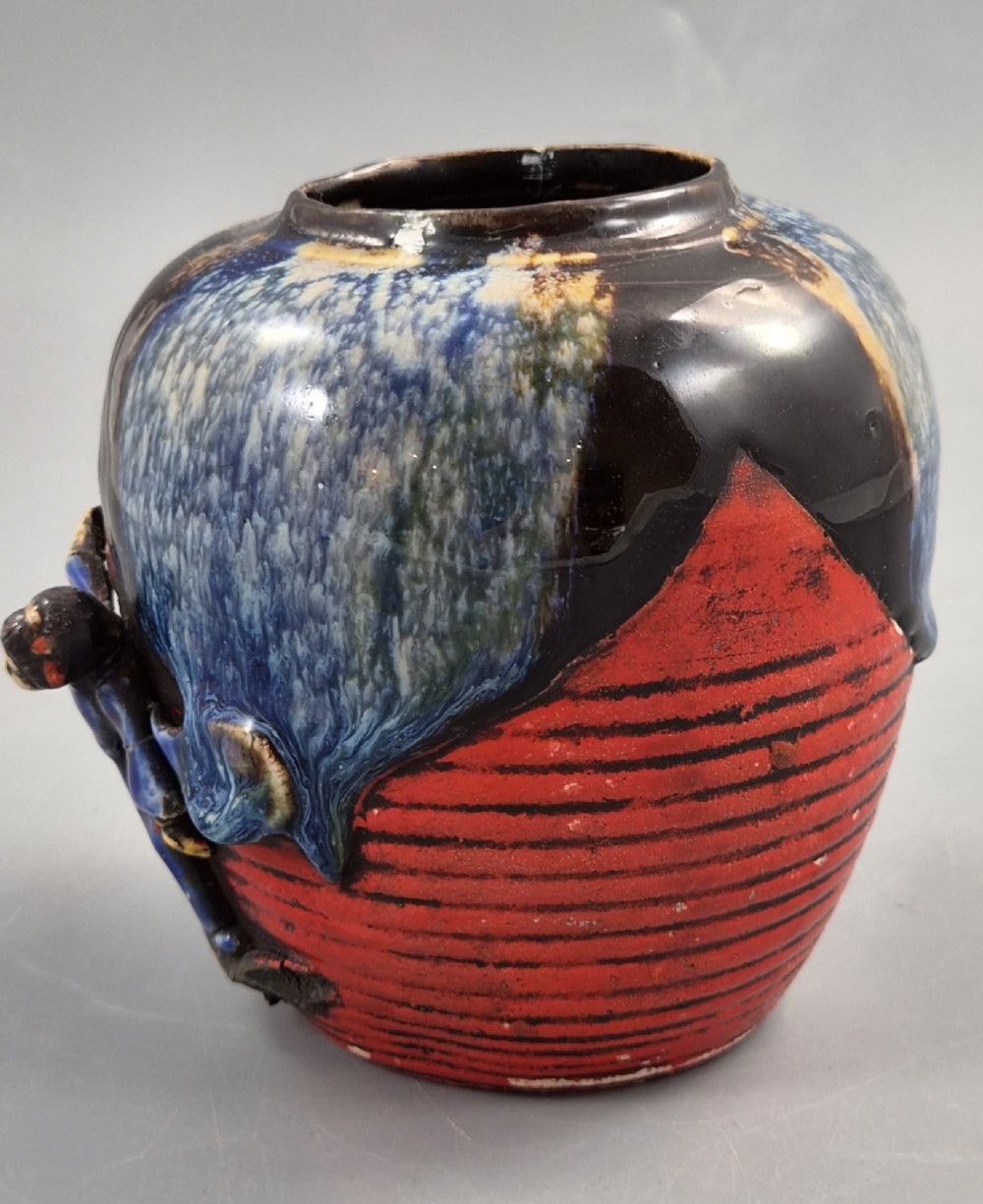 A rounded Japanese Sumida Gawa ceramic jar featuring heavy blue flambe dripping glaze over a ribbed red body with an applied relief climbing figure. View II