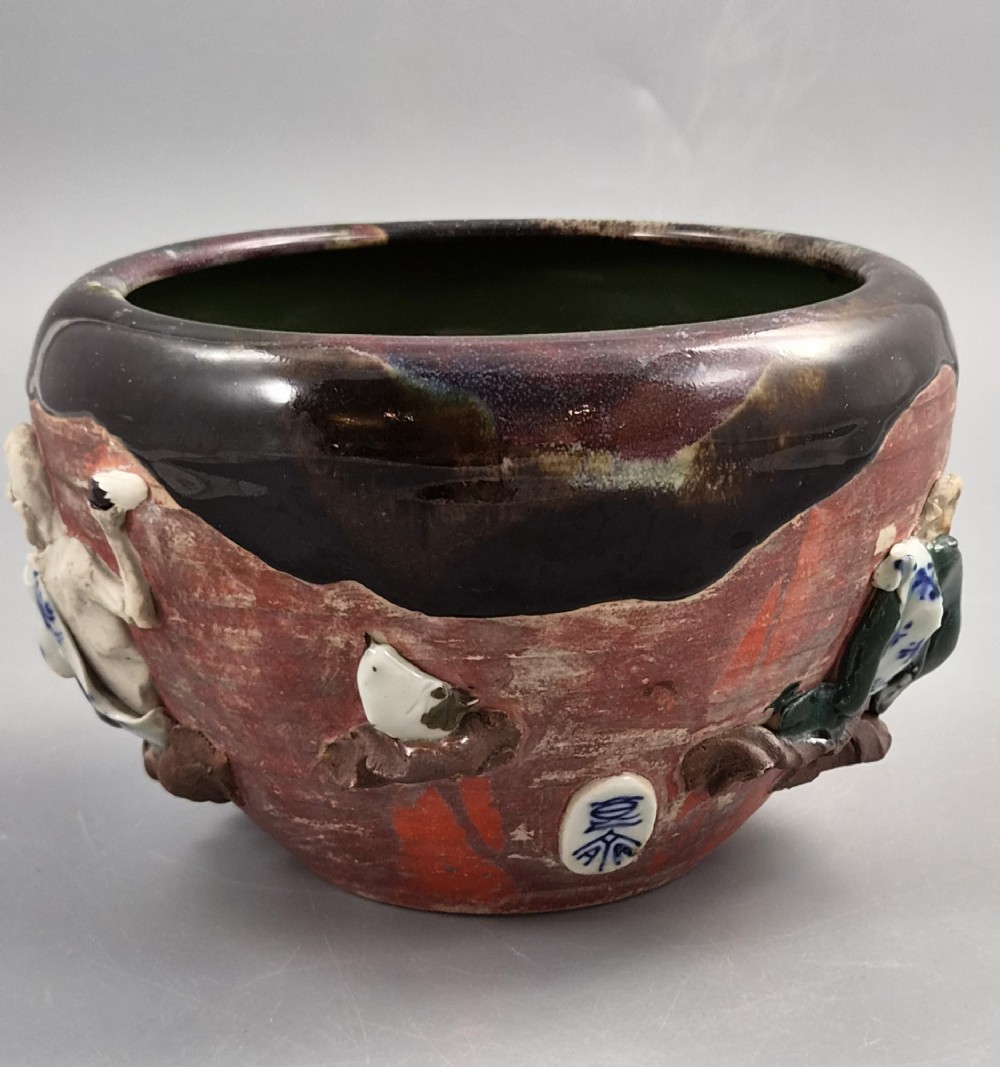 An antique Japanese Sumida Gawa ceramic bowl featuring heavy dark flambe glaze over a red terracotta body adorned with three-dimensional applied figures in relief. View III