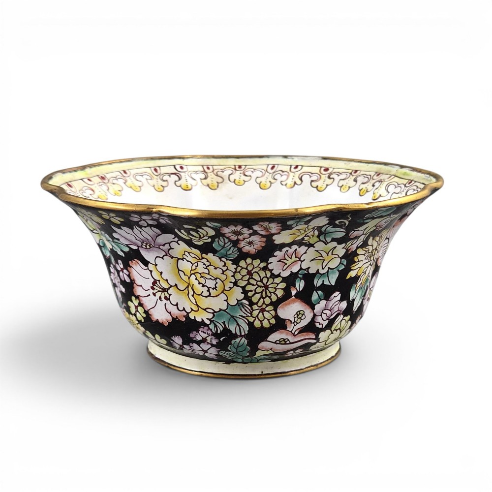 A Chinese Canton enamel-on-copper bowl featuring a lobed rim, a black ground exterior decorated with colourful floral motifs, and a white interior with a patterned border.