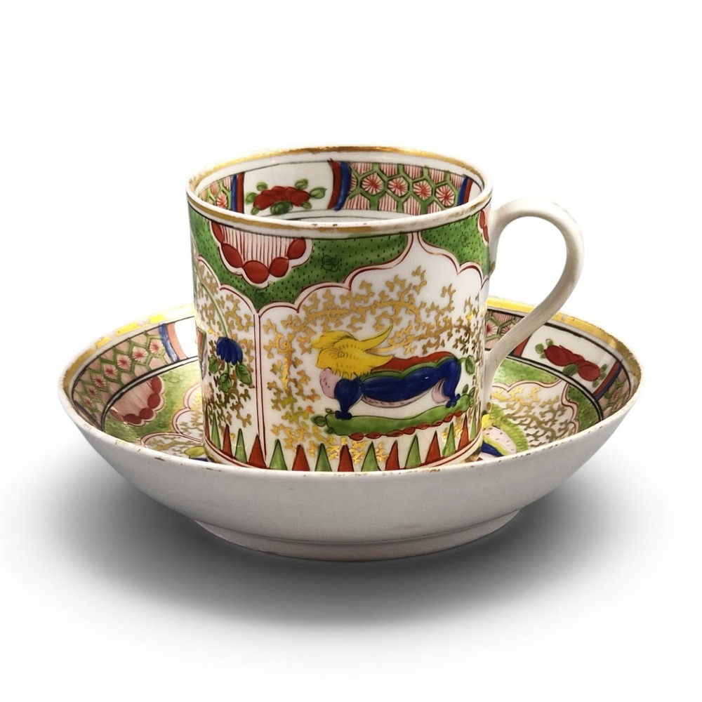 A Regency period Worcester porcelain coffee cup and matching saucer decorated in the hand-painted Dragon in Compartments pattern with green, red, blue, and gilt detailing.