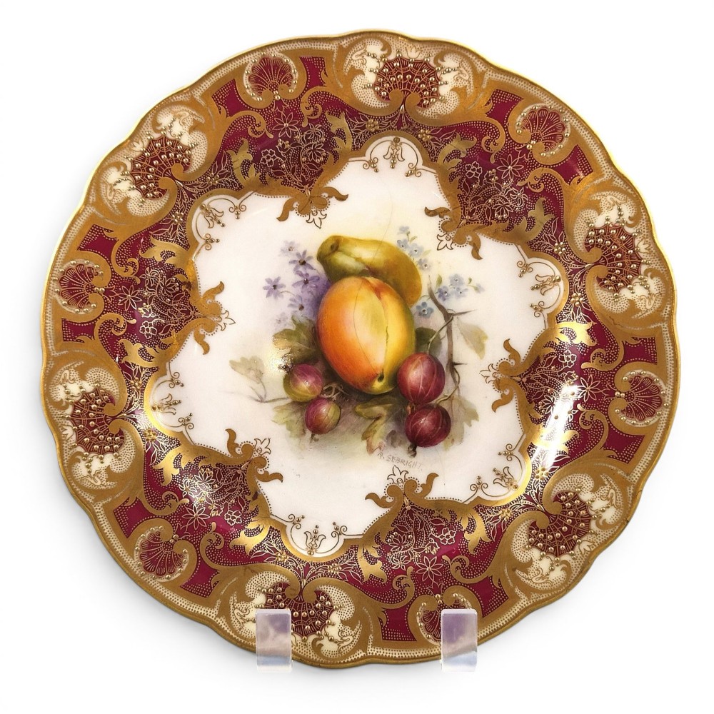 A circular porcelain cabinet plate featuring a central hand-painted fruit still life surrounded by a heavily gilded burgundy rim with scalloped edges.