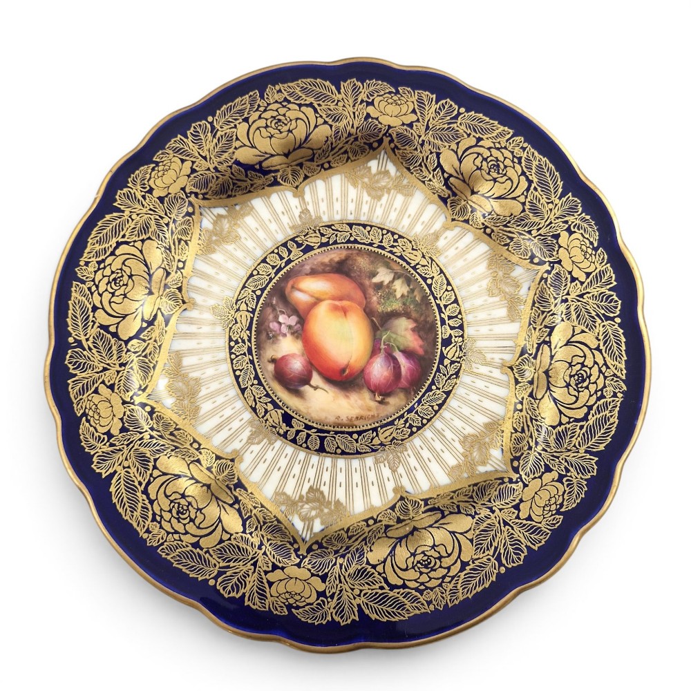 A circular porcelain cabinet plate featuring a central hand-painted fruit still life surrounded by a gilded sunburst pattern and a cobalt blue rim embellished with gold floral scrollwork.