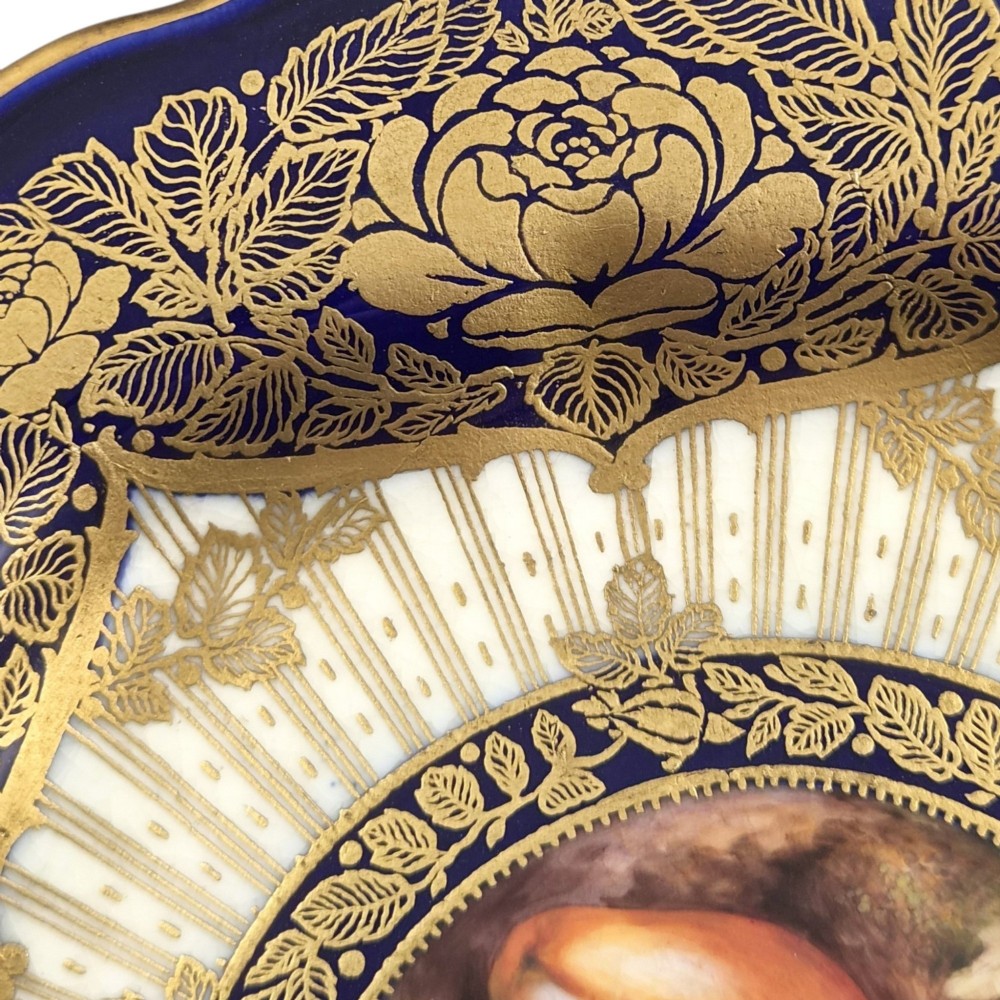 A circular porcelain cabinet plate featuring a central hand-painted fruit still life surrounded by a gilded sunburst pattern and a cobalt blue rim embellished with gold floral scrollwork. View IV