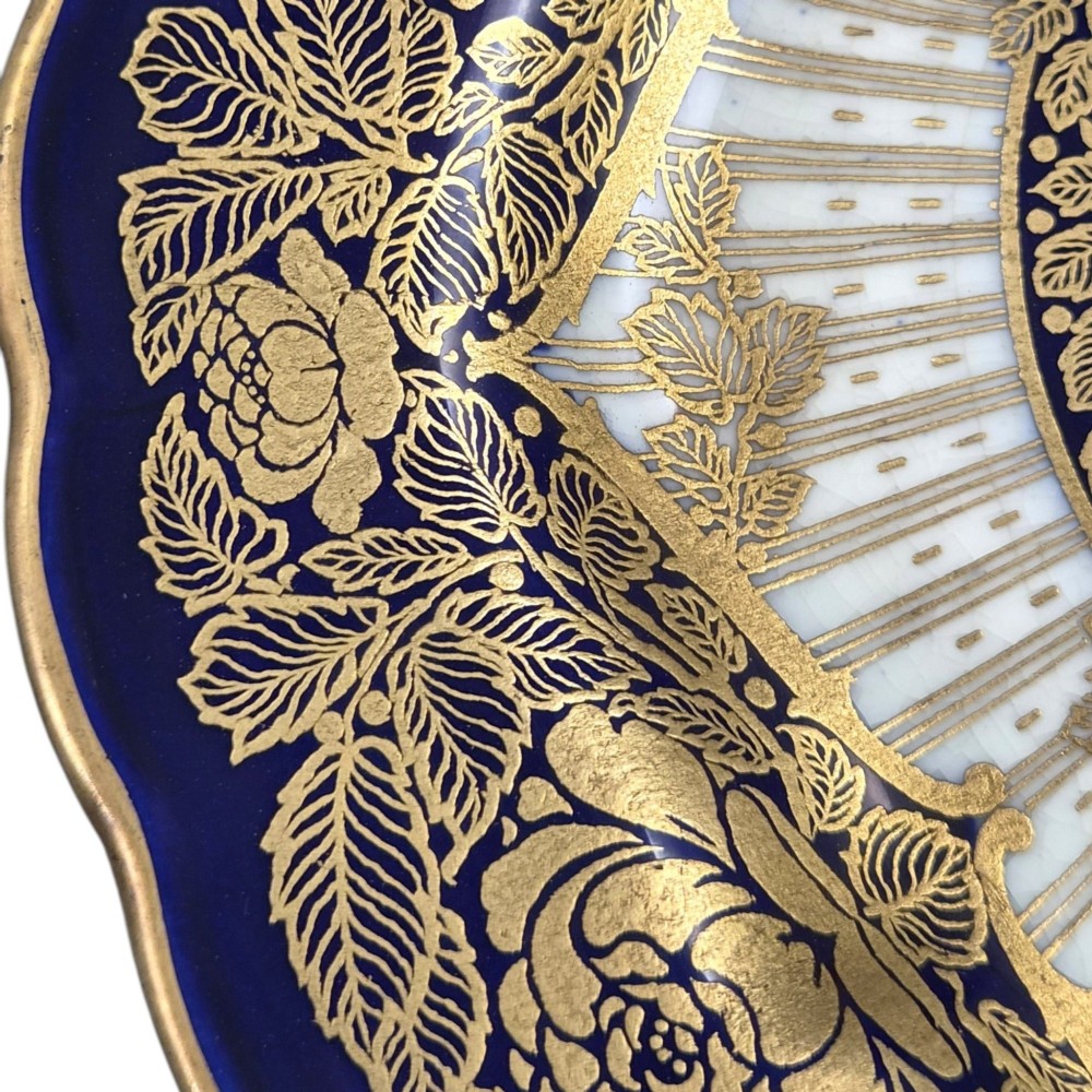 A circular porcelain cabinet plate featuring a central hand-painted fruit still life surrounded by a gilded sunburst pattern and a cobalt blue rim embellished with gold floral scrollwork. View V