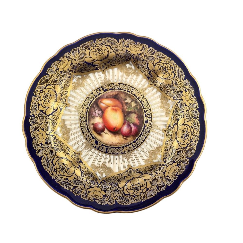 A circular porcelain cabinet plate featuring a central hand-painted fruit still life surrounded by a gilded sunburst pattern and a cobalt blue rim embellished with gold floral scrollwork. View X