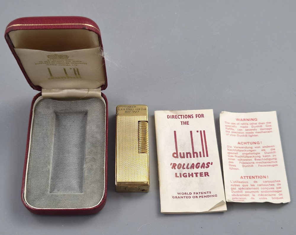A 1967 gold-plated Dunhill Rollagas pocket lighter with a textured barley pattern, displayed alongside its fitted red presentation case and original printed instruction booklet.