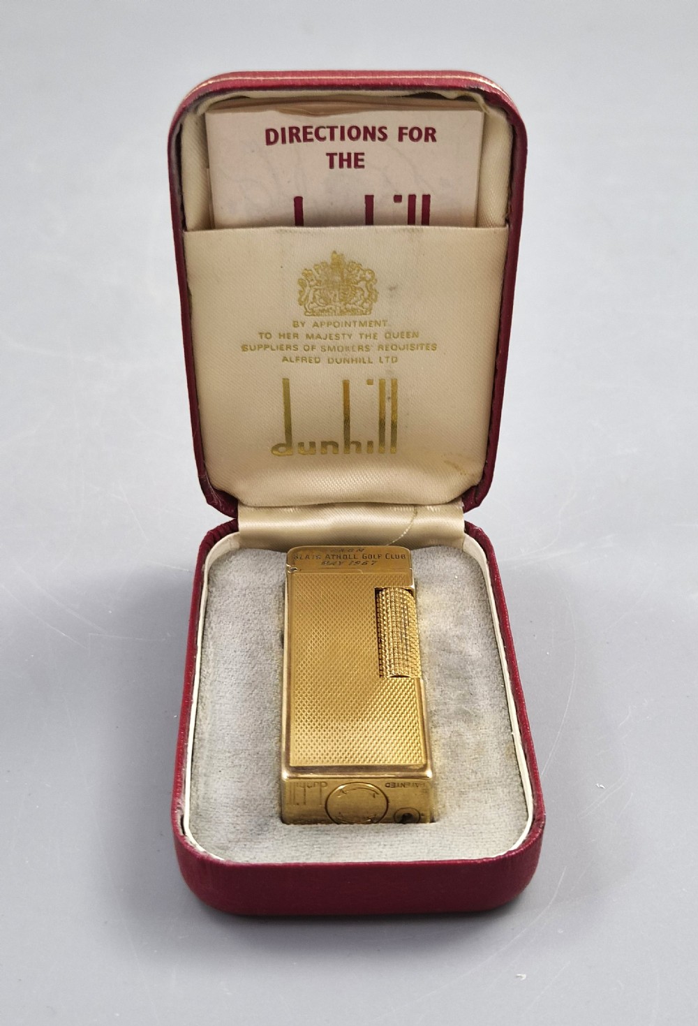 A 1967 gold-plated Dunhill Rollagas pocket lighter with a textured barley pattern, displayed alongside its fitted red presentation case and original printed instruction booklet. View II