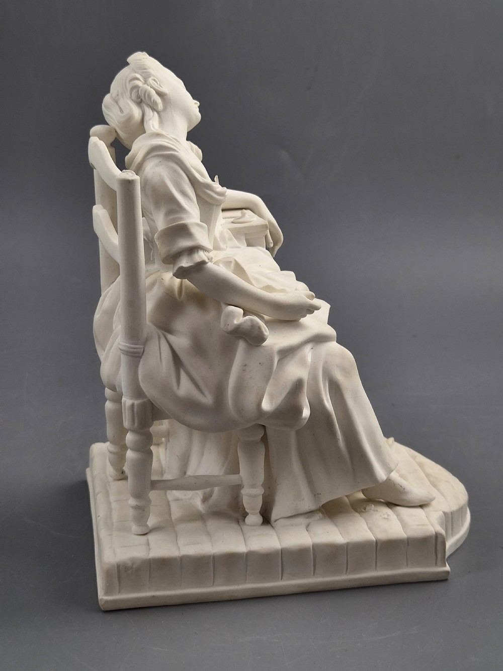 A 19th-century off-white Parian porcelain figure depicting a sleeping woman seated in a ladder-back chair on a rectangular base.
