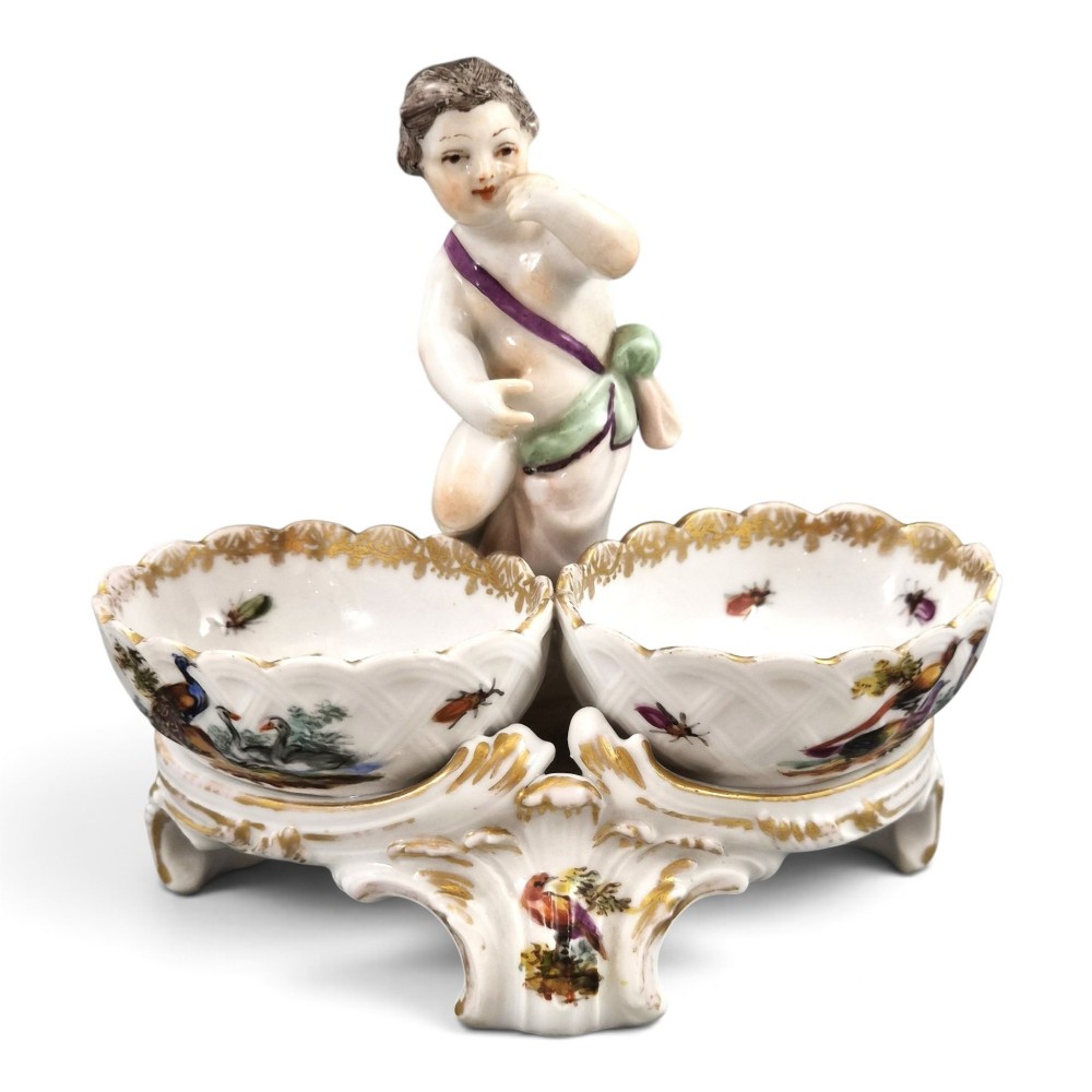 A 19th-century German hard-paste porcelain double salt cellar featuring a central sculpted figure of a cherub flanked by two shell-shaped bowls hand-painted with birds and insects.