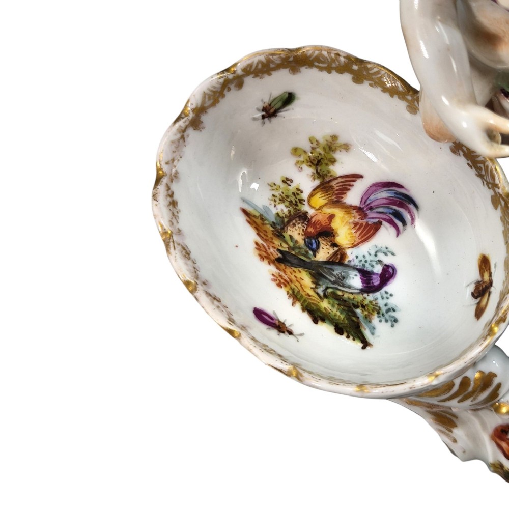 A 19th-century German hard-paste porcelain double salt cellar featuring a central sculpted figure of a cherub flanked by two shell-shaped bowls hand-painted with birds and insects. View IX