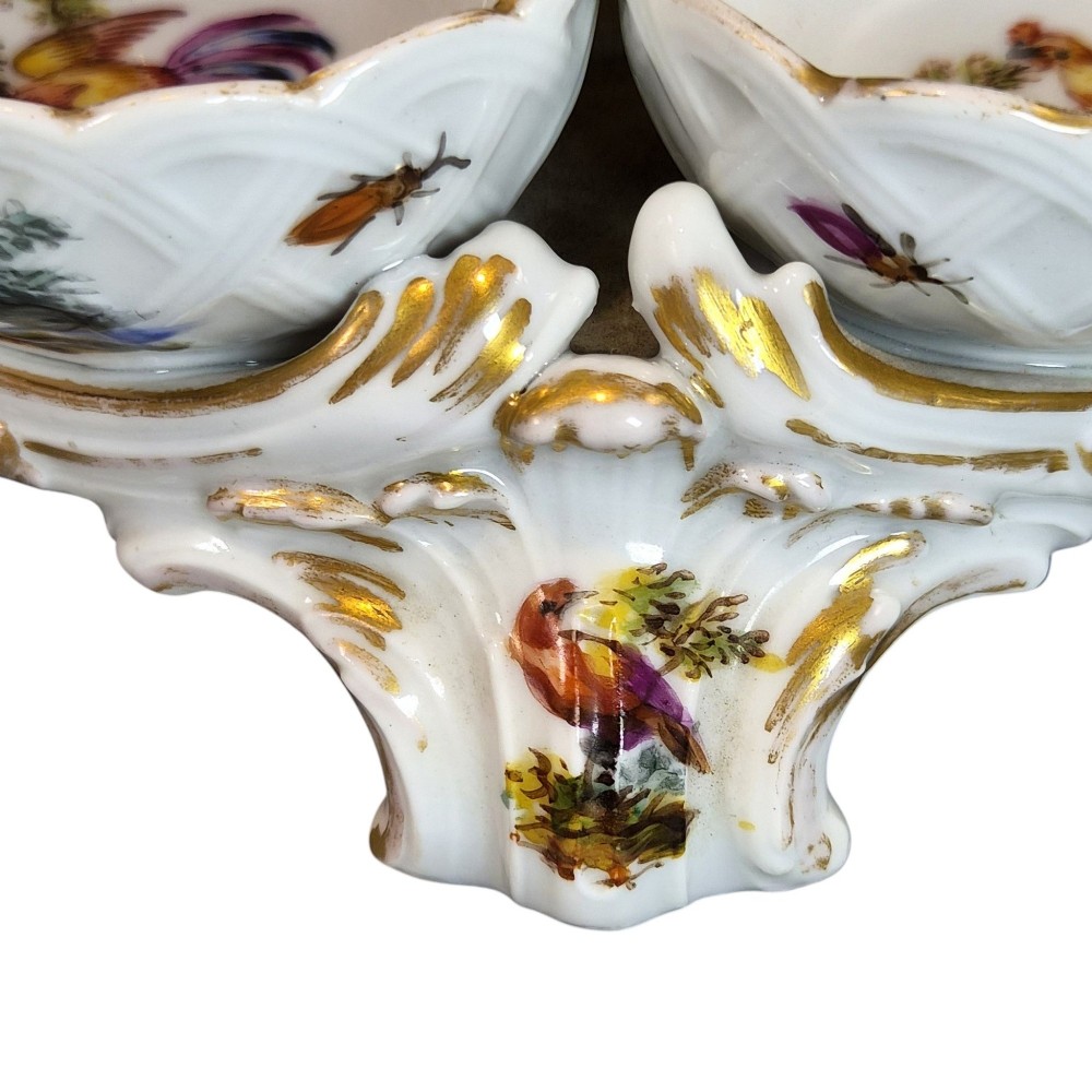 A 19th-century German hard-paste porcelain double salt cellar featuring a central sculpted figure of a cherub flanked by two shell-shaped bowls hand-painted with birds and insects. View X