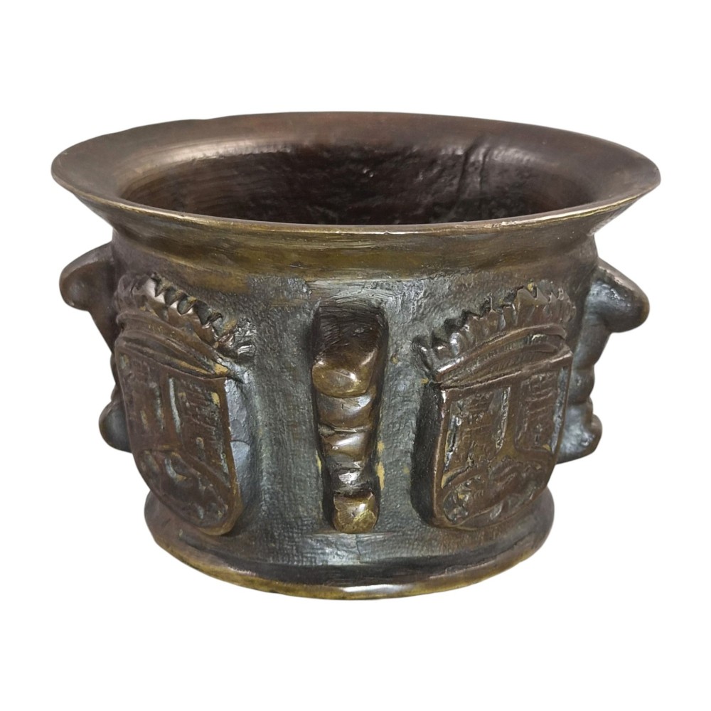 A heavy 17th-century cast bronze apothecary mortar featuring raised heraldic coat of arms motifs and side lugs around a flared rim. View XI