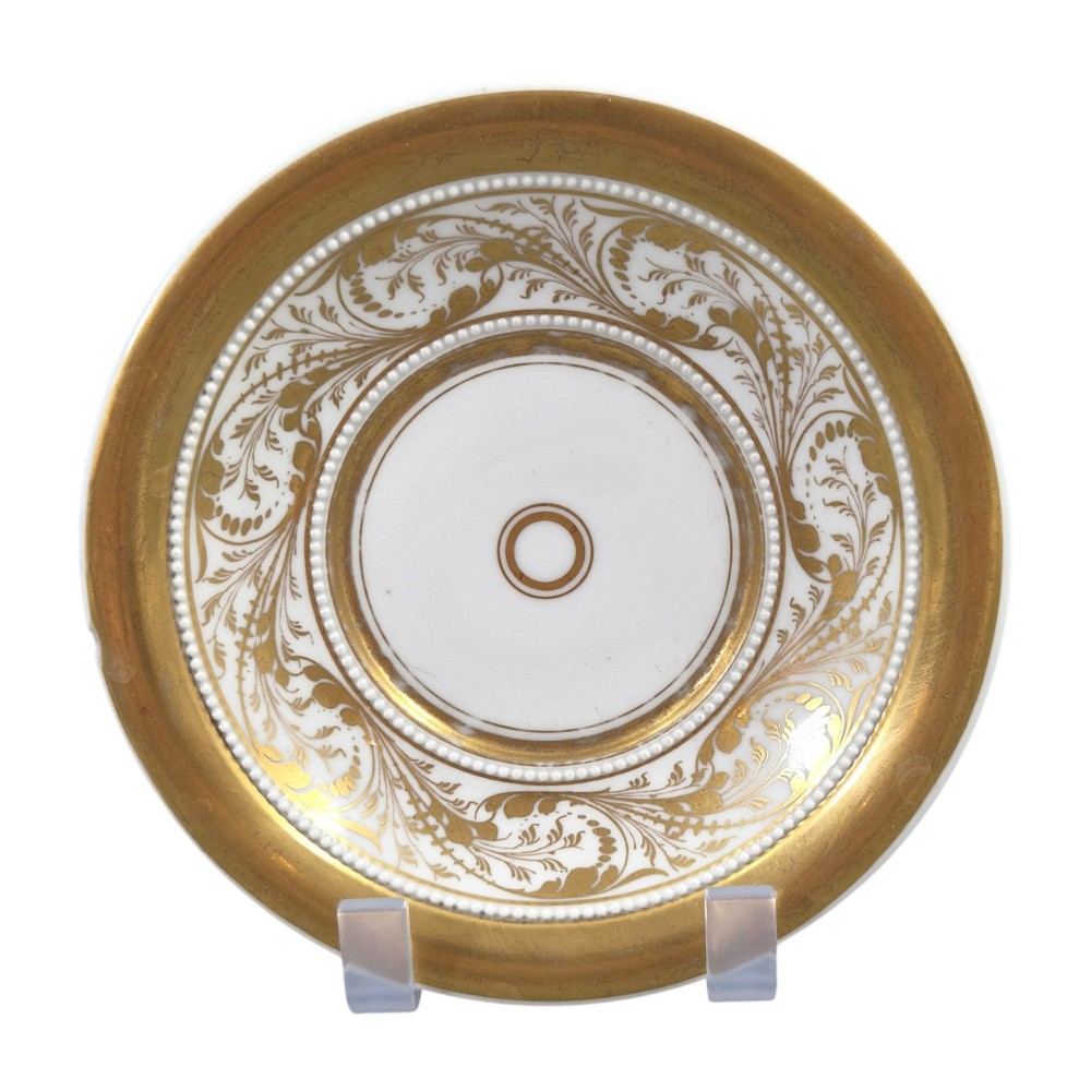 An antique white and gilt porcelain presentation cup with an elevated loop handle sitting on a matching gilded saucer. View II
