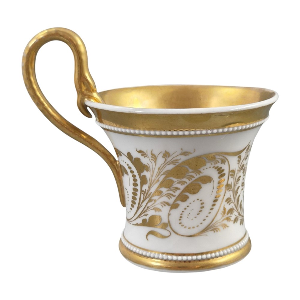 An antique white and gilt porcelain presentation cup with an elevated loop handle sitting on a matching gilded saucer. View VIII
